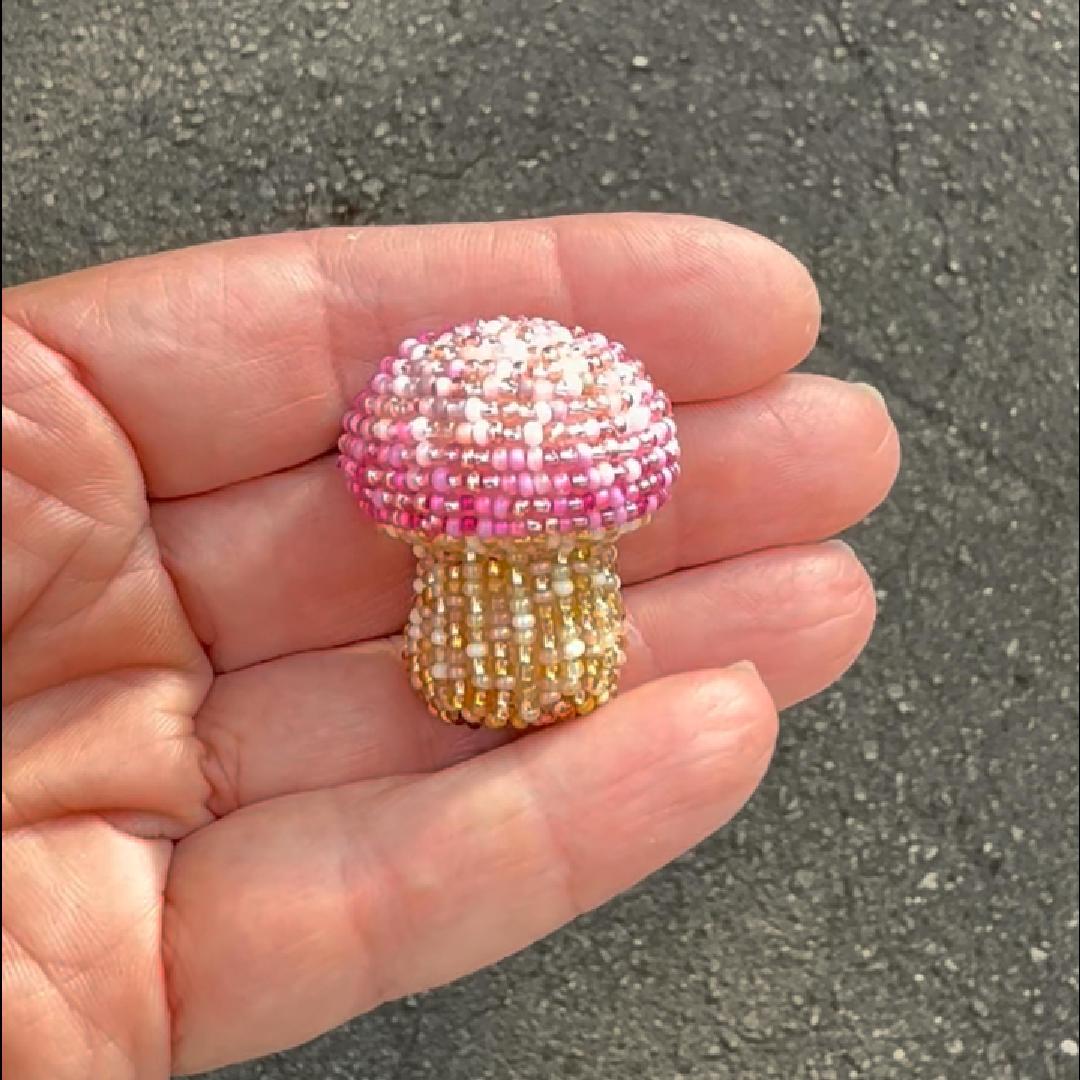 A small mushroom-shaped beaded brooch held in a person's hand, with a pink and white beaded cap and a gold beaded stem, against an outdoor asphalt background.