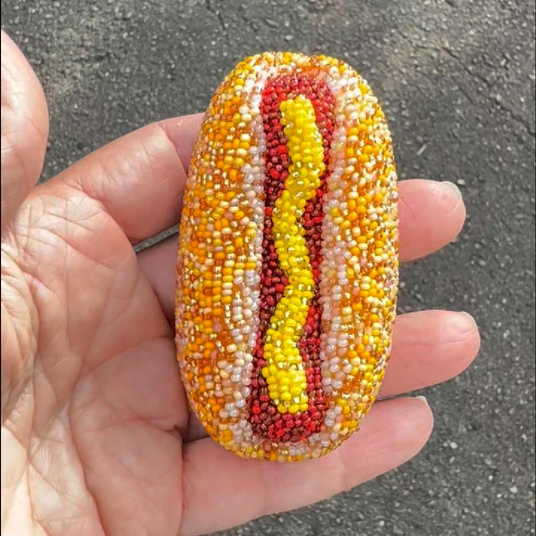 A hand holding a hot dog-shaped brooch made of many small beads, with a yellow center, red hot dog, and white and beige sides, against an outdoor gray ground background.