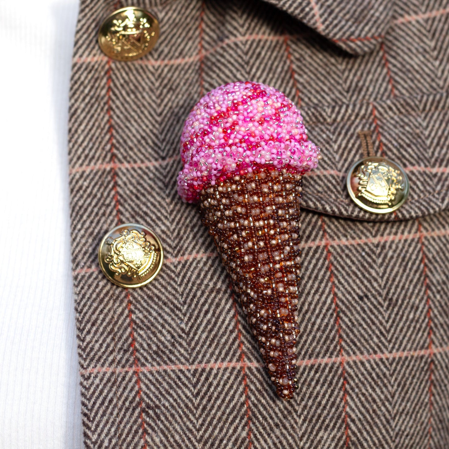 Closeup of a handmade beaded pink ice cream cone brooch on a brown textured jacket with gold buttons.