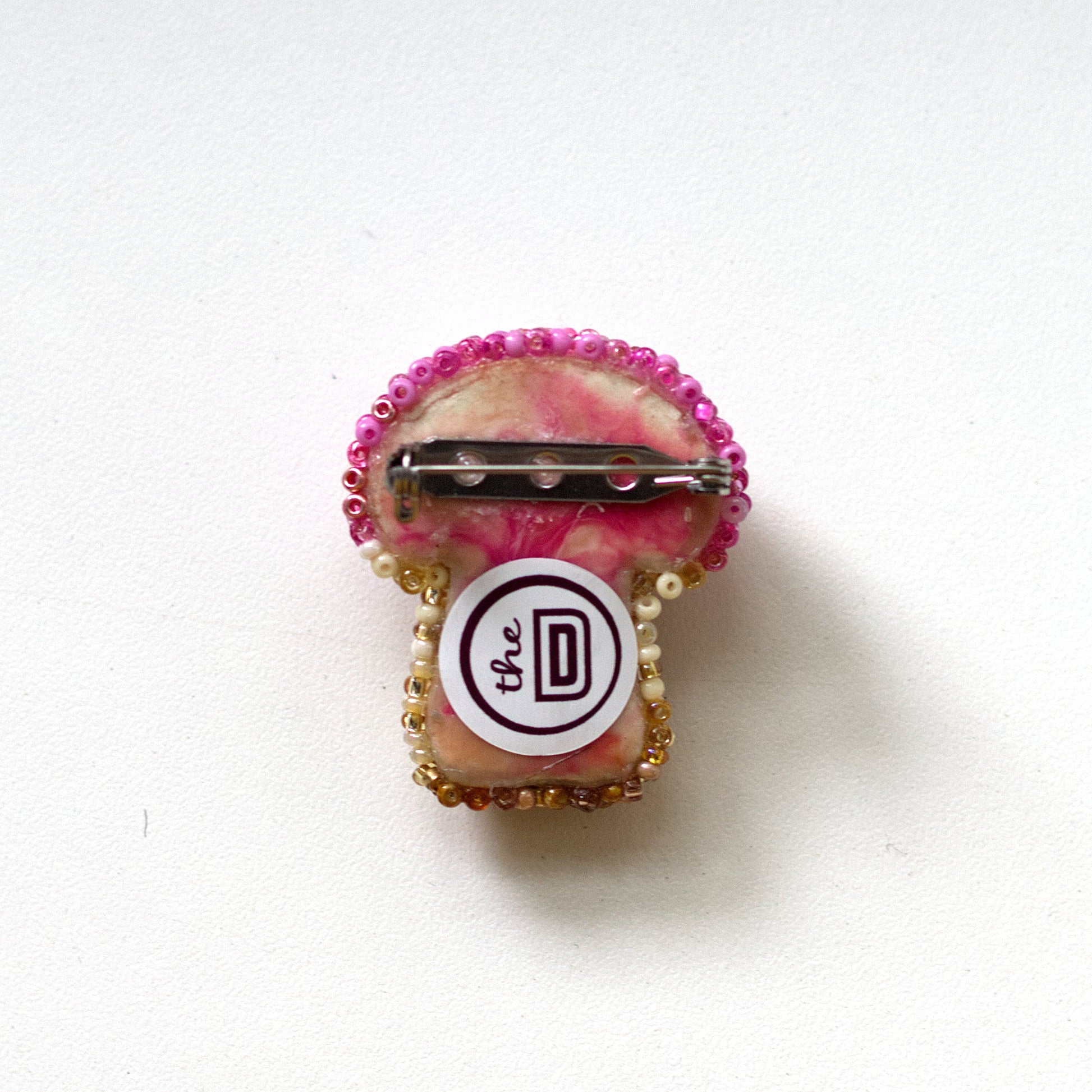 The back of a small beaded brooch with a pink marbled top, gold beaded edges, and a circular white center featuring a pin and branding, photographed on a plain white background.