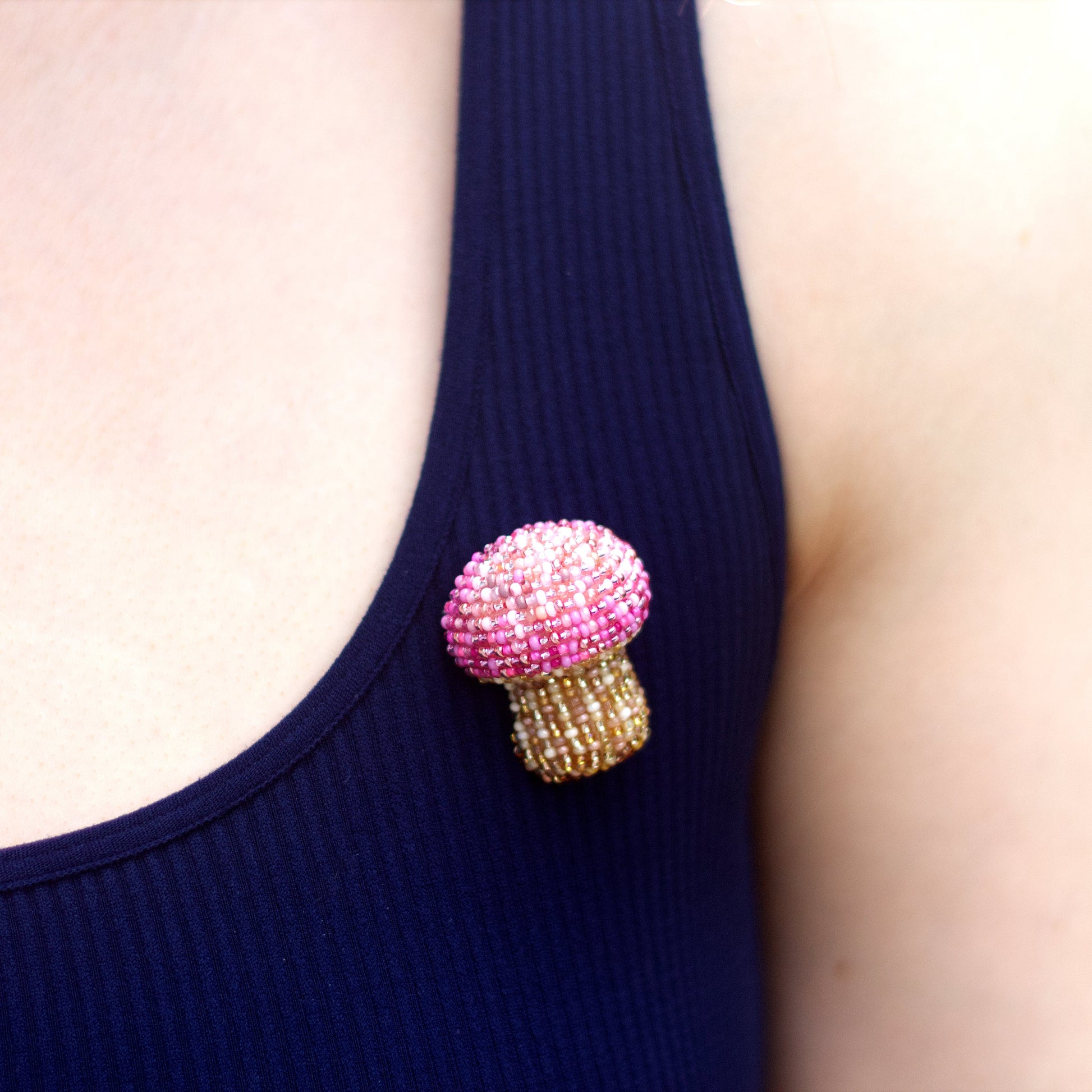 A close-up of a person wearing a mushroom-shaped beaded brooch with a pink crown and a gold textured stem, pinned to the chest of a navy ribbed tank top.