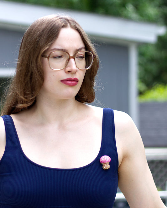 A woman with glasses and brown hair wearing a navy blue ribbed tank top, featuring a small pink mushroom-shaped brooch pinned to the chest, set against a blurred outdoor background of a gray house and green foliage.