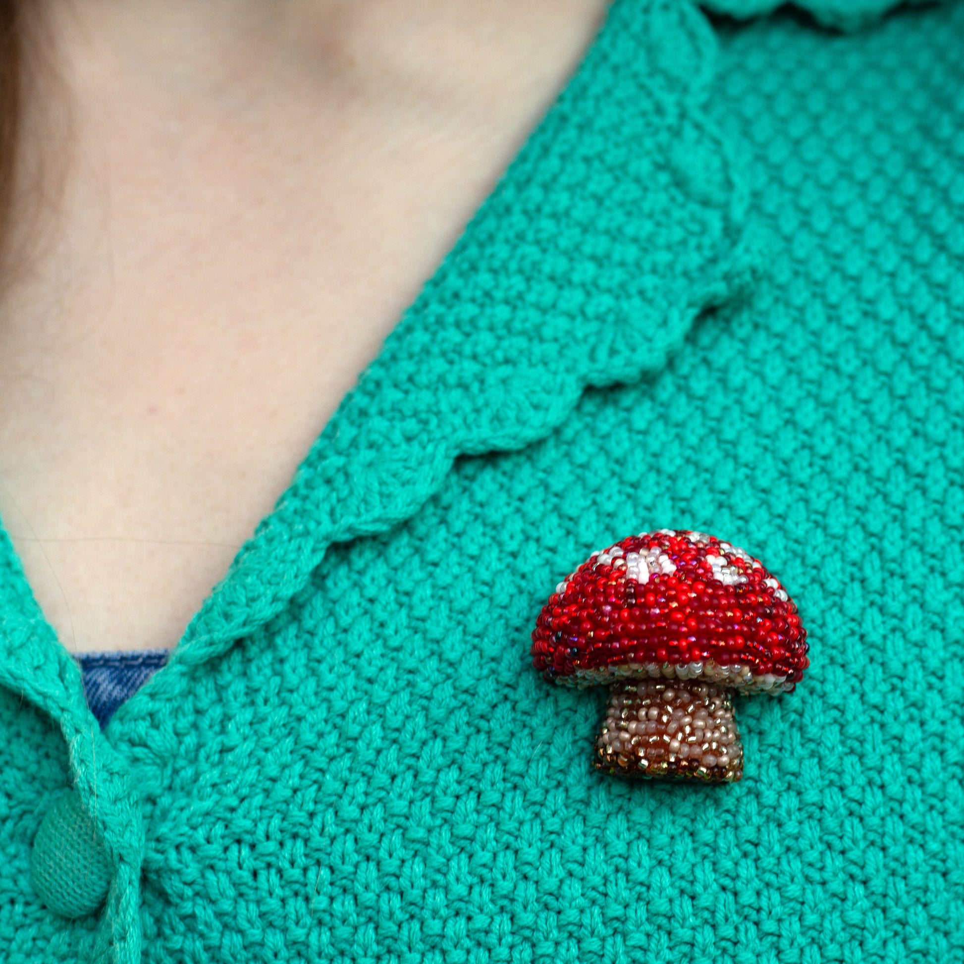Closeup view of a red and white handmade beaded mushroom brooch worn on a green cardigan.