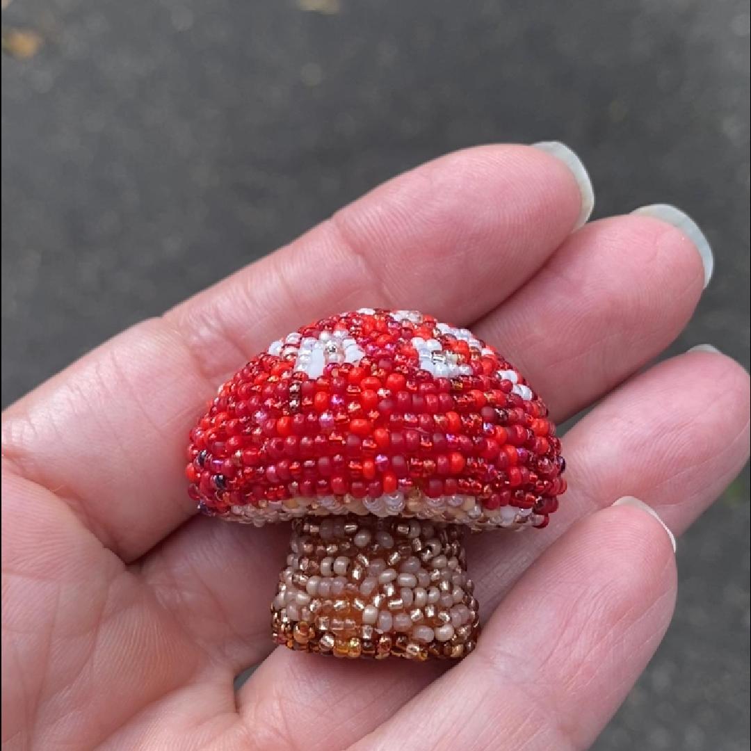 Hand holding a red and white handmade beaded mushroom brooch  in the sunlight.