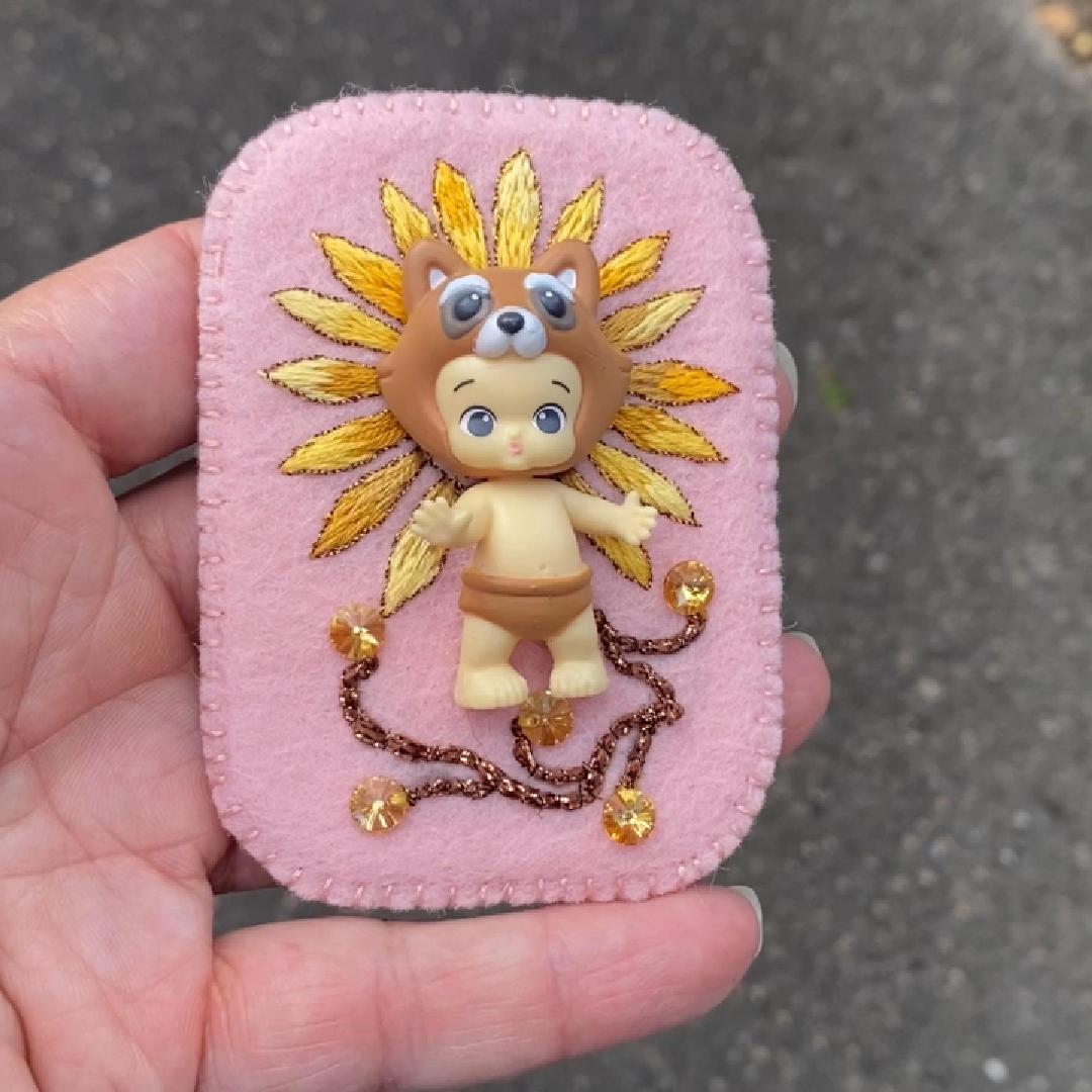 Hand holding a daisy embroidered handmade brooch with Happi Twozie in the sunlight.