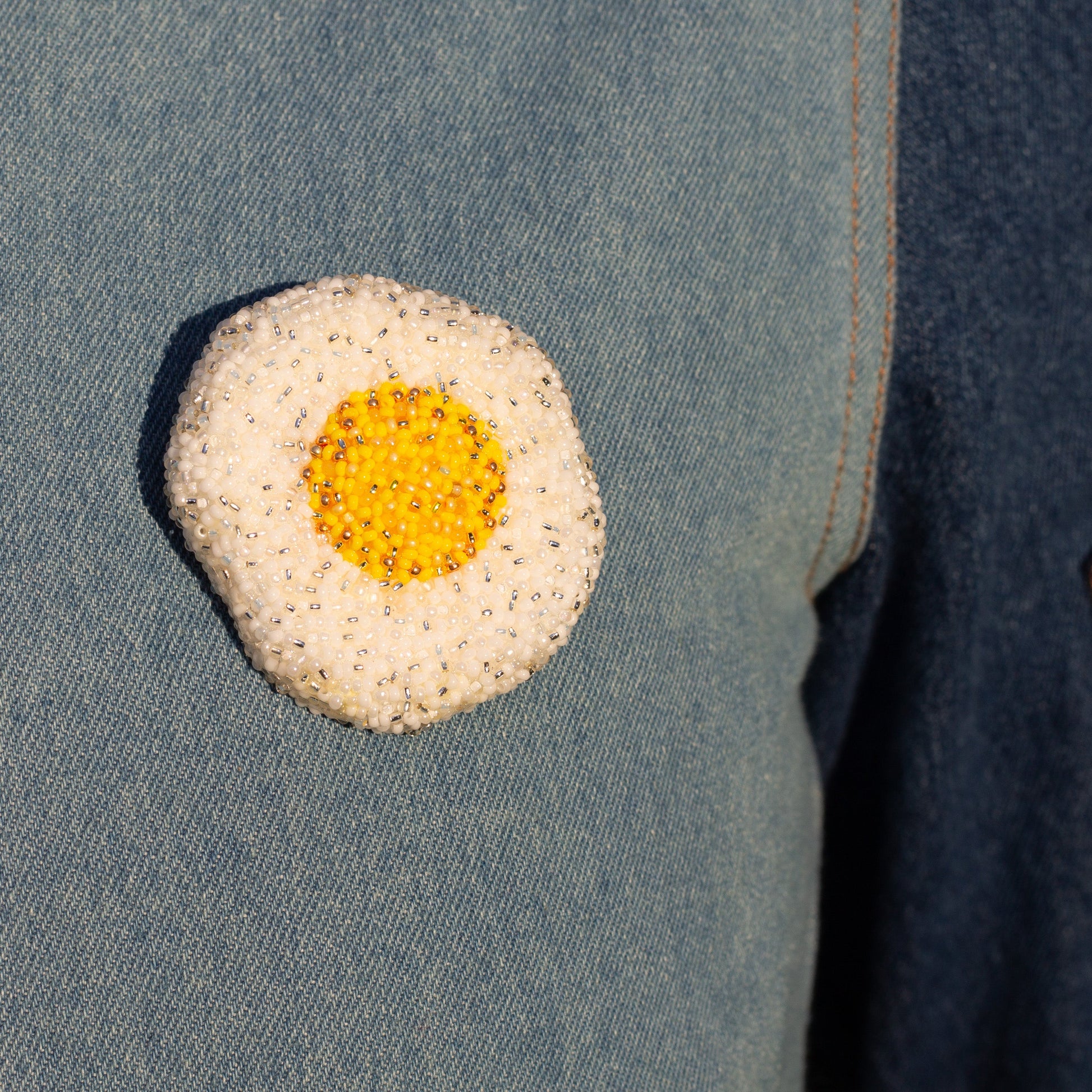 Closeup of a handmade beaded fried egg brooch on a denim jacket.