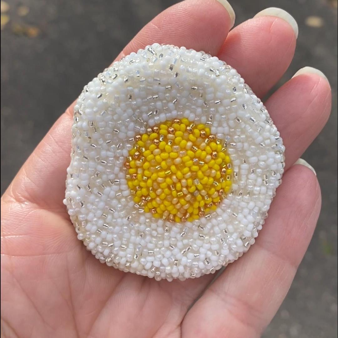 A hand holding a beaded fried egg brooch in the sunlight.