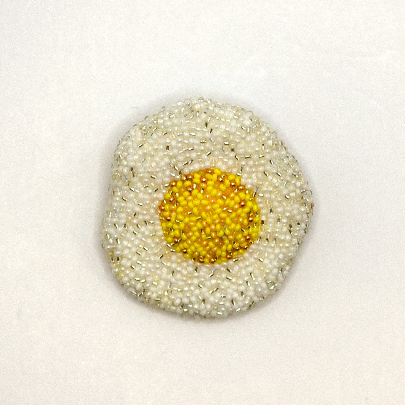 A handmade beaded fried egg brooch on a white background.