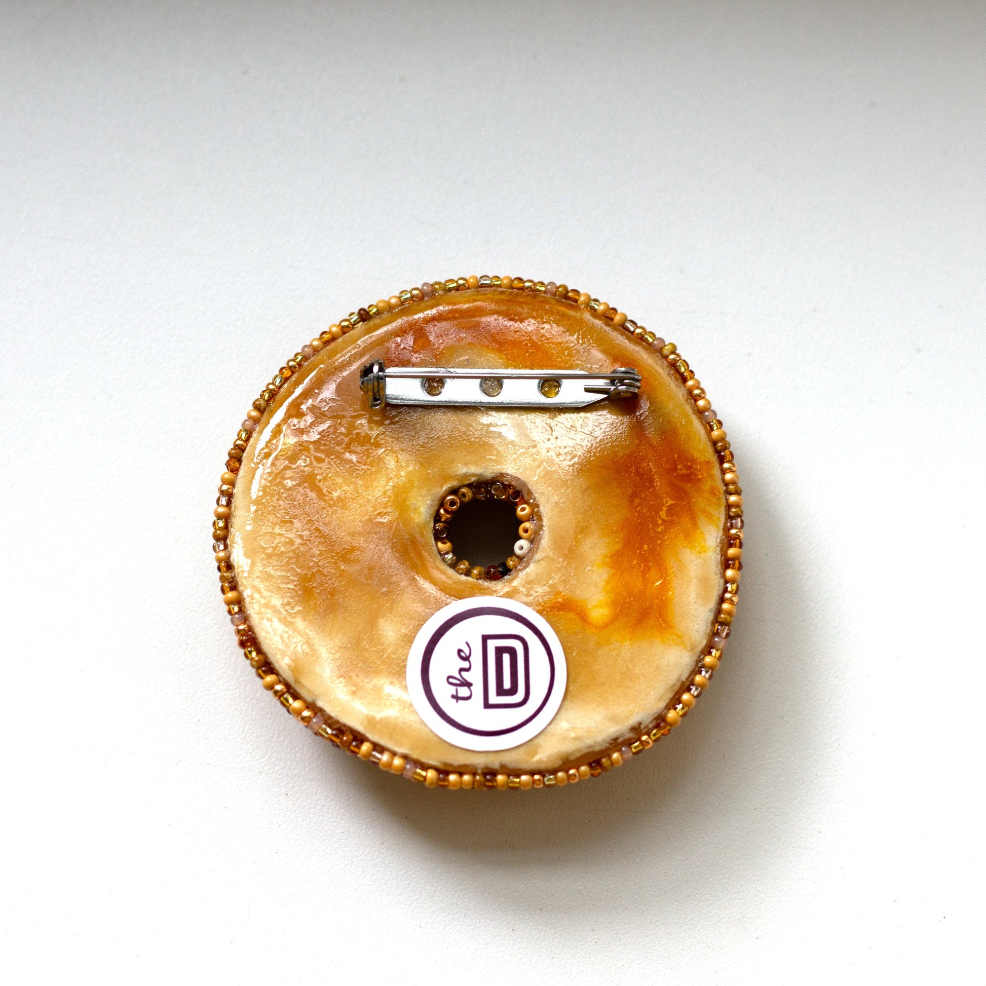 The back of a beaded everything bagel brooch with a gold beaded edge and a metal pin back, featuring a white circular sticker with the brand name 'the DD' on a plain white background.