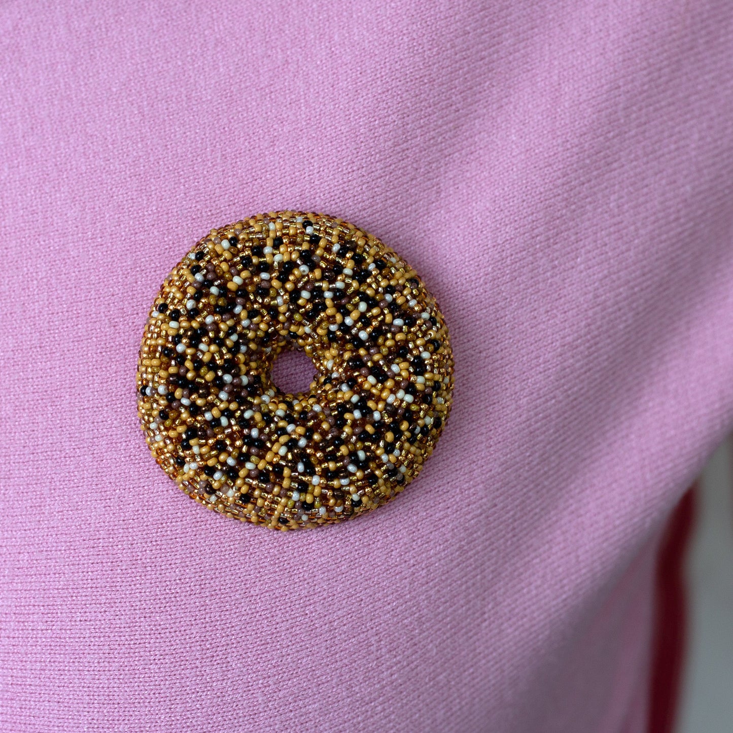 A beaded everything bagel brooch covered in gold, black, and white seed beads, pinned onto a light pink textured fabric.