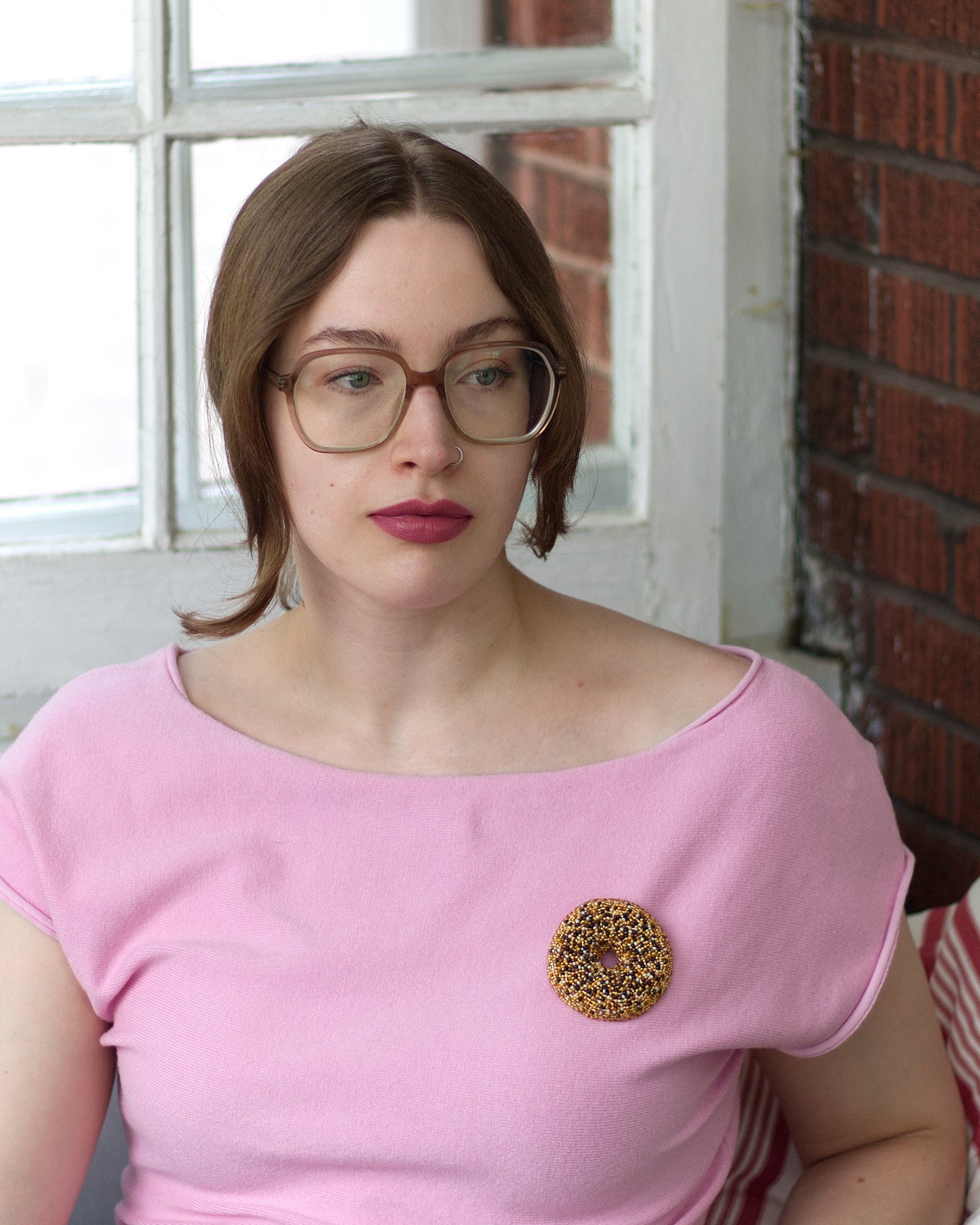 A woman with glasses and brown hair sits indoors wearing a pink boat-neck t-shirt with an everything bagel beaded brooch on the chest, positioned in front of a brick wall and window.