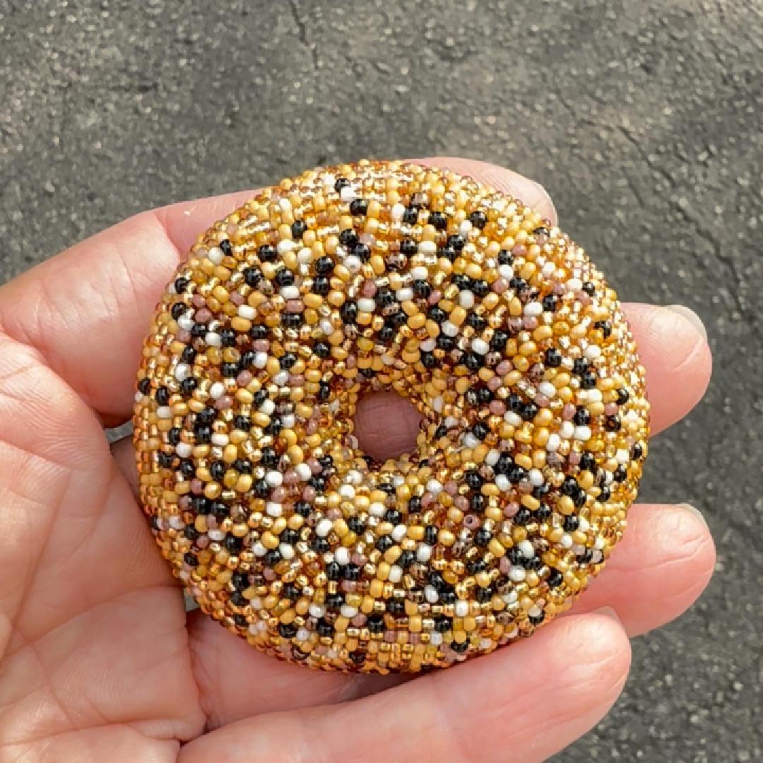 A hand holding a beaded everything bagel brooch covered in gold, black, and white seed beads, photographed outdoors against a gray paved surface.