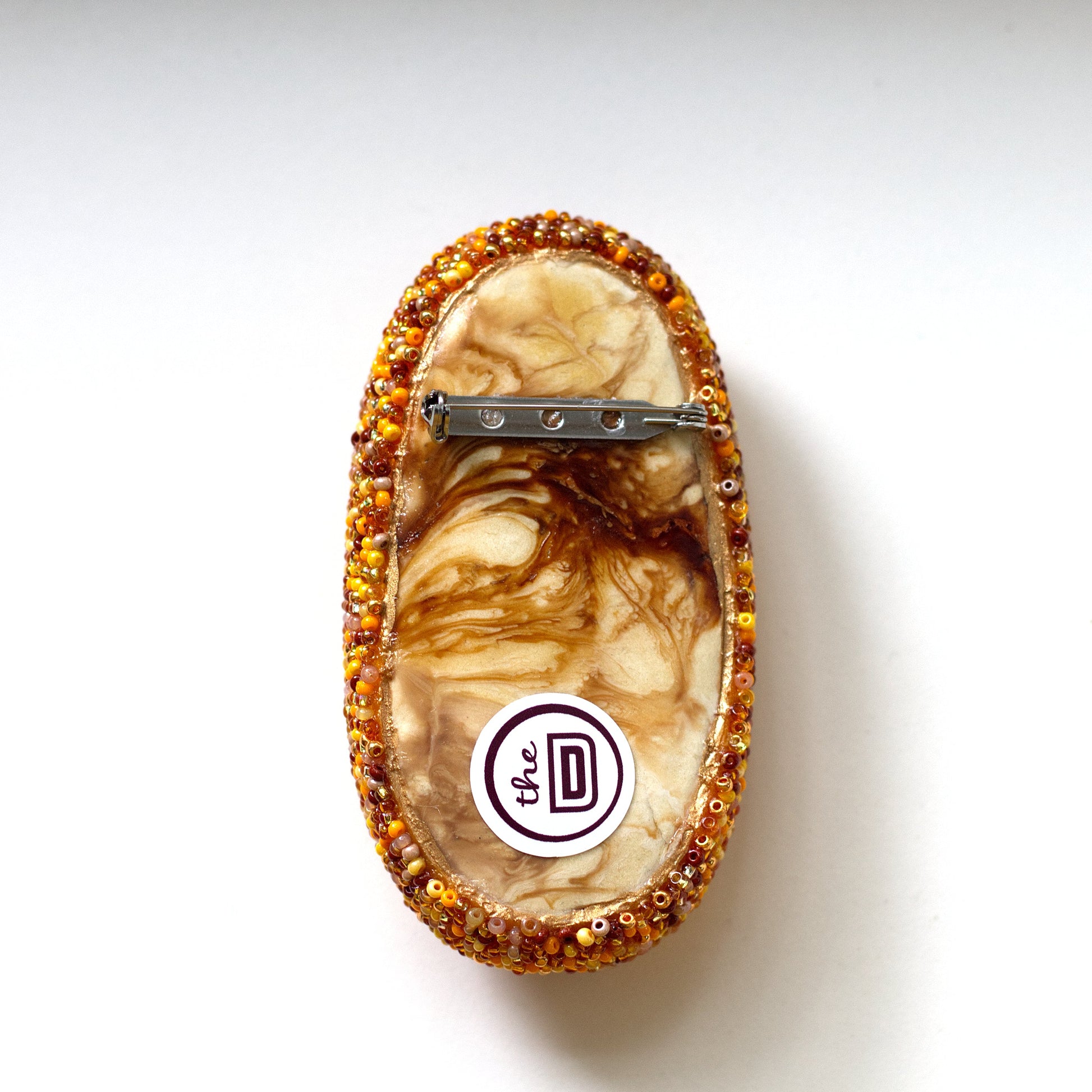 An oval handmade brooch back with a marbled brown and cream pattern, bordered by sparkling amber-orange beads, featuring a metal pin back and a circular brand sticker.