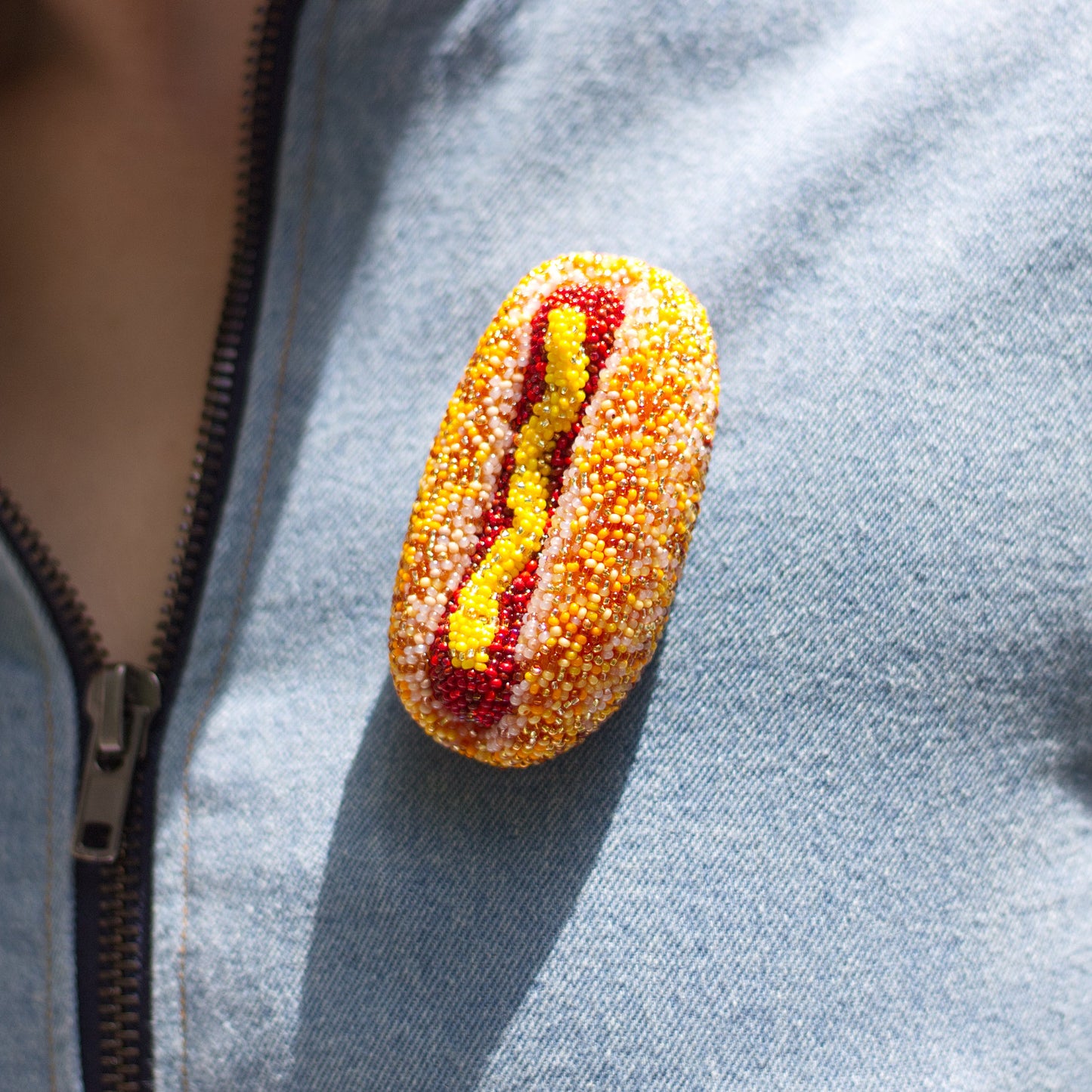 A handmade hot dog-shaped brooch made of yellow and red beads, pinned onto the chest of a light blue denim vest.