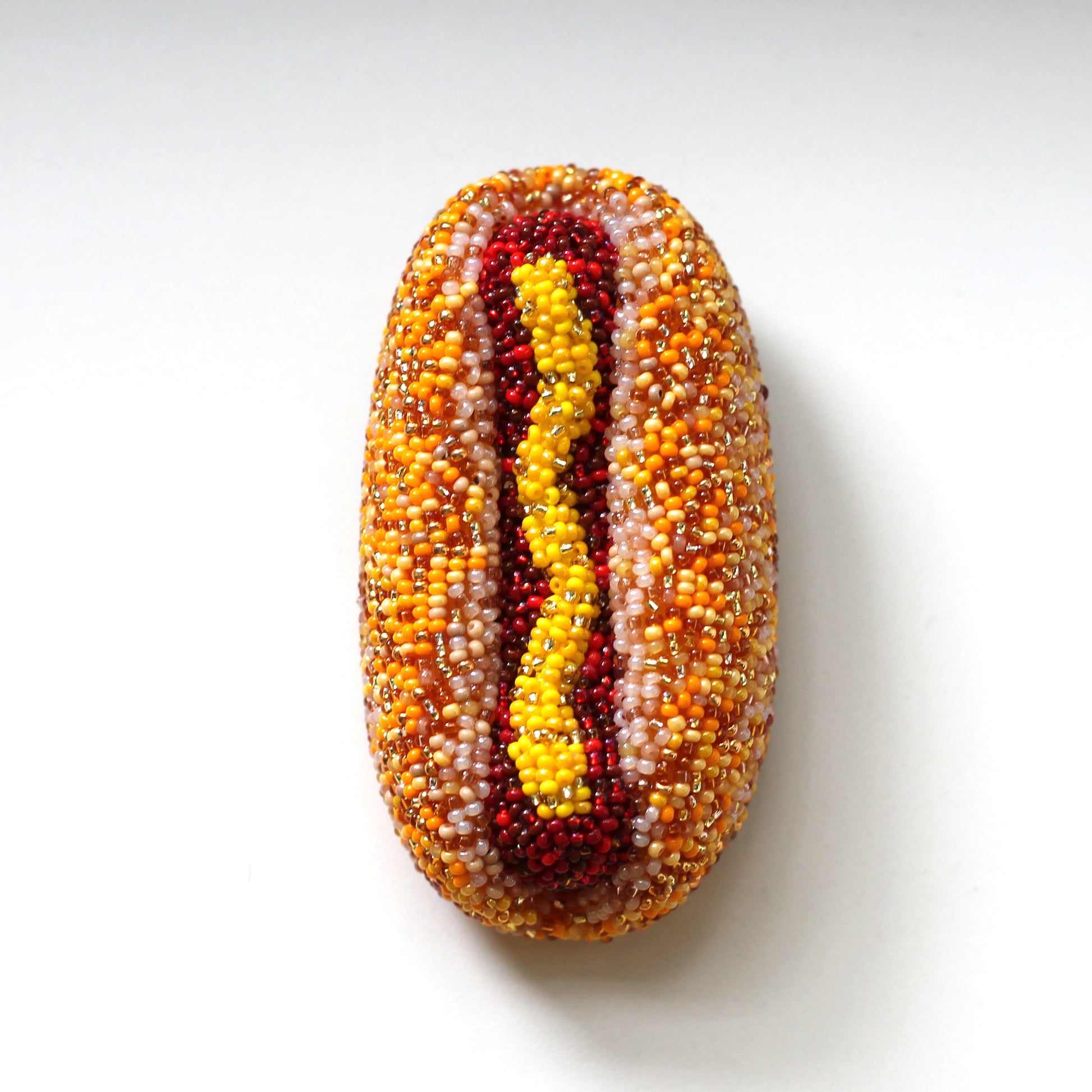 A handmade hot dog brooch made from small seed-like beads, featuring a tan and beige bun with a wavy yellow mustard line and red hot dog detail, centered on a plain white background.