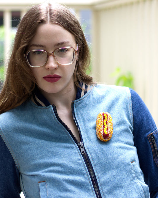 A woman with long light brown hair and glasses wearing a light blue and dark blue knit bomber jacket with a beaded hot dog pin on the chest, standing in a bright indoor setting.