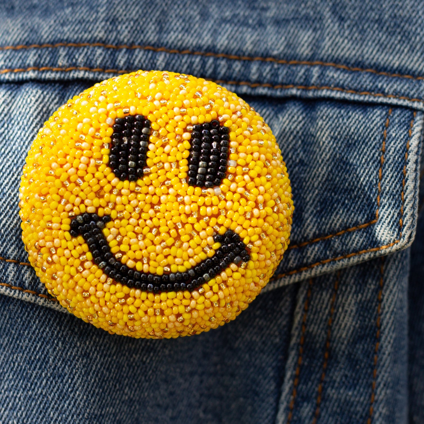 Closeup of a yellow smiley face handmade beaded brooch worn on a faded denim jacket.