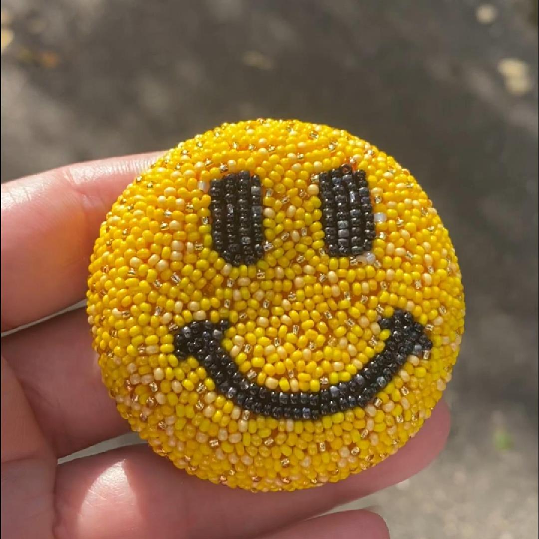 Hand holding a yellow smiley face beaded brooch in the sunlight.