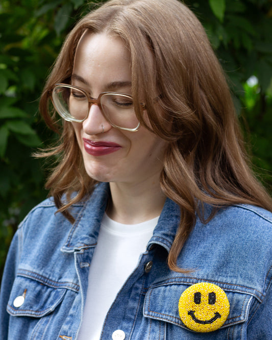 A yellow smiley face handmade beaded brooch worn on a faded denim jacket by a young woman.