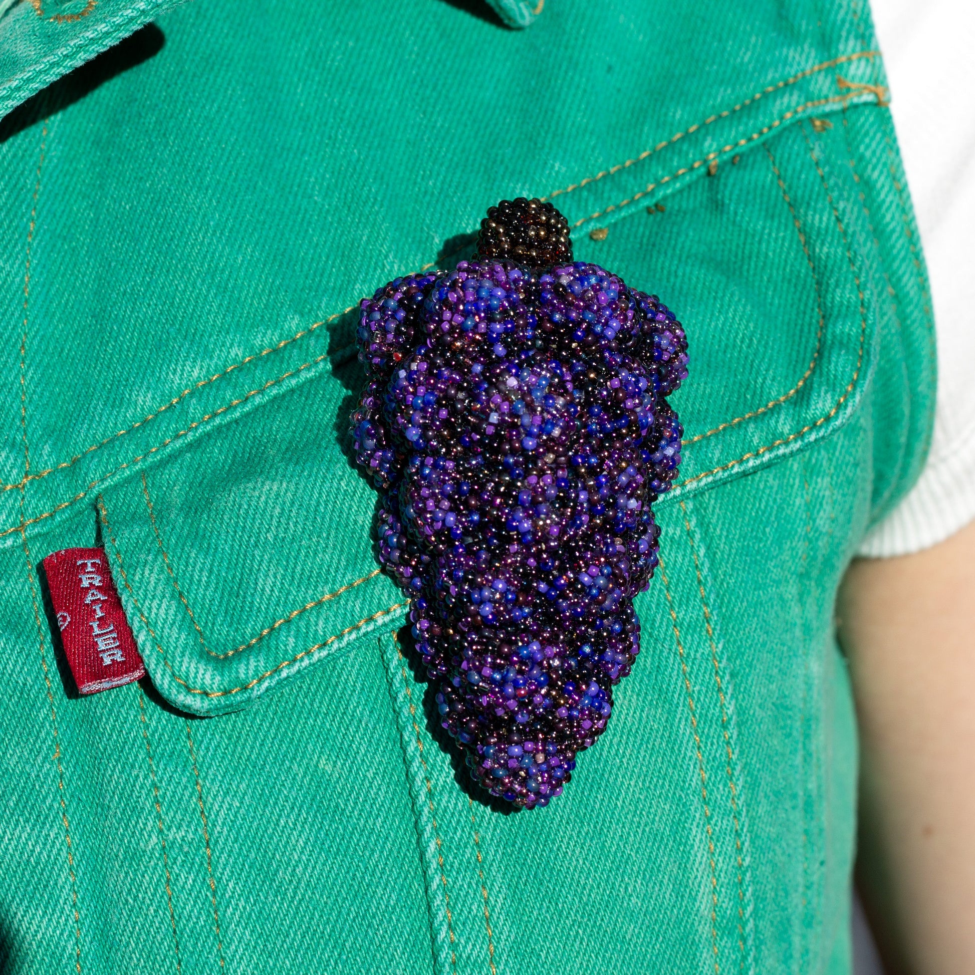 Closeup of a purple handmade beaded grape bunch brooch worn on a green vest pocket.