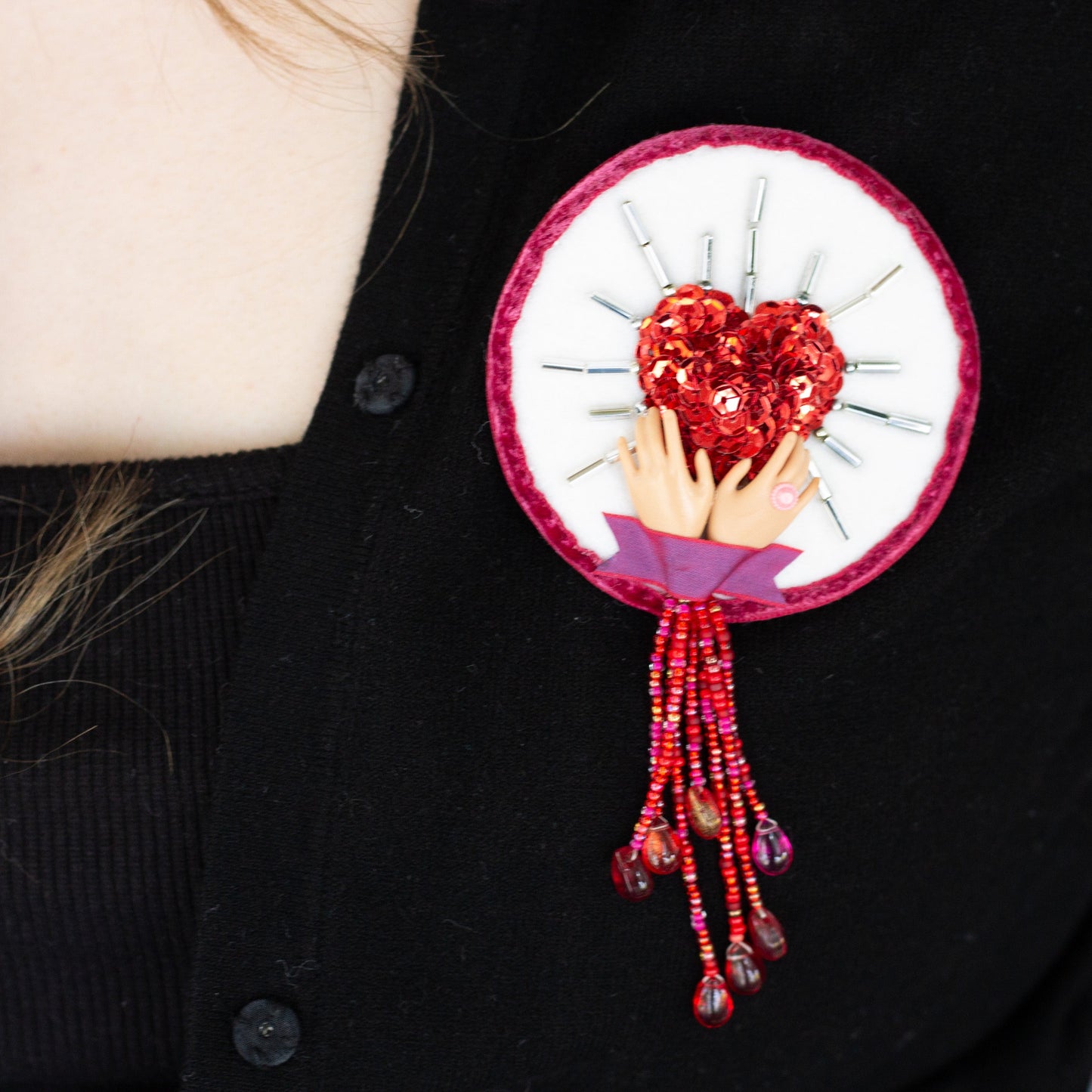 Closeup of a handmade brooch featuring a sequined heart cradled by doll hands worn on a black sweater.
