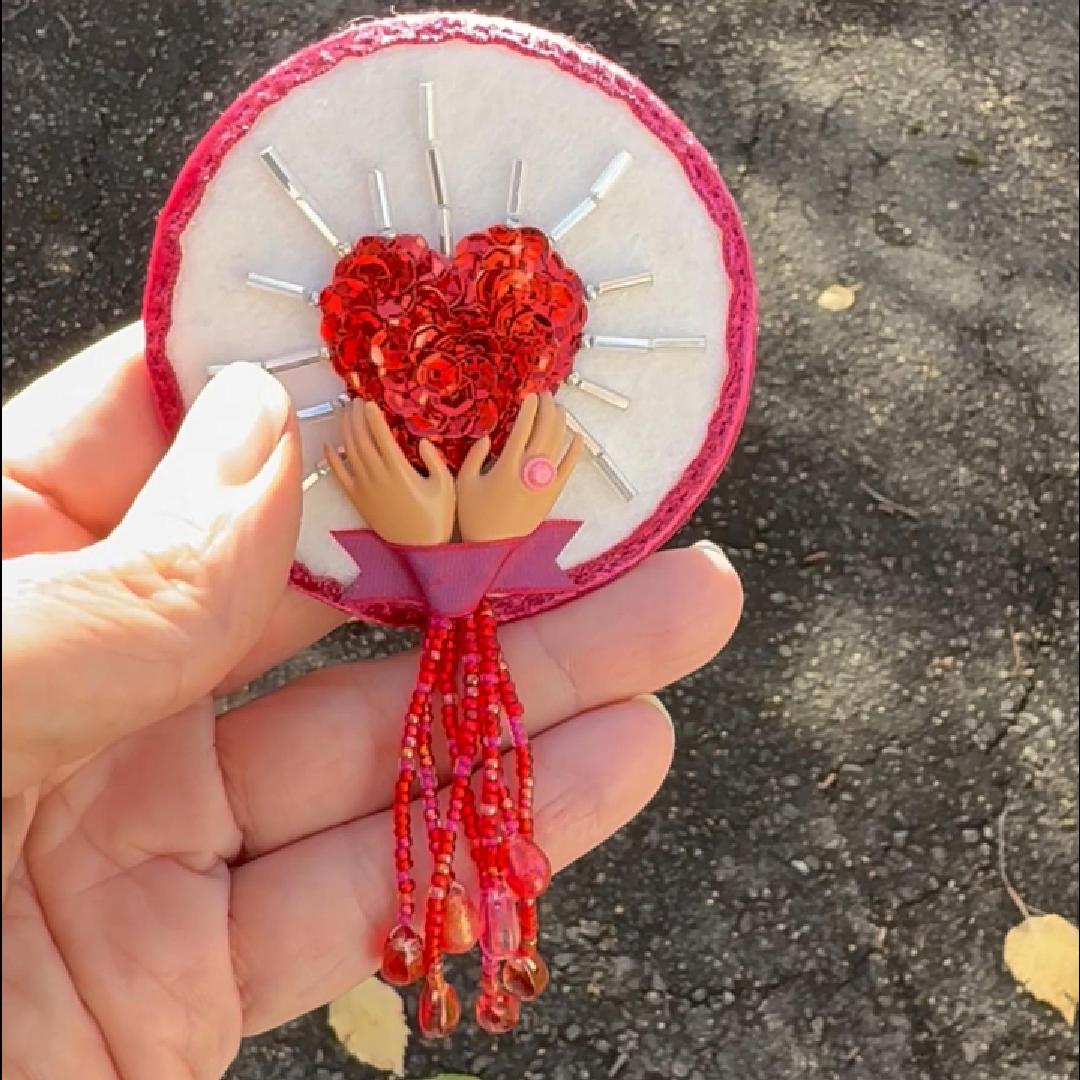 Hand holding a brooch featuring a sequined heart cradled by doll hands shown in the sunlight.