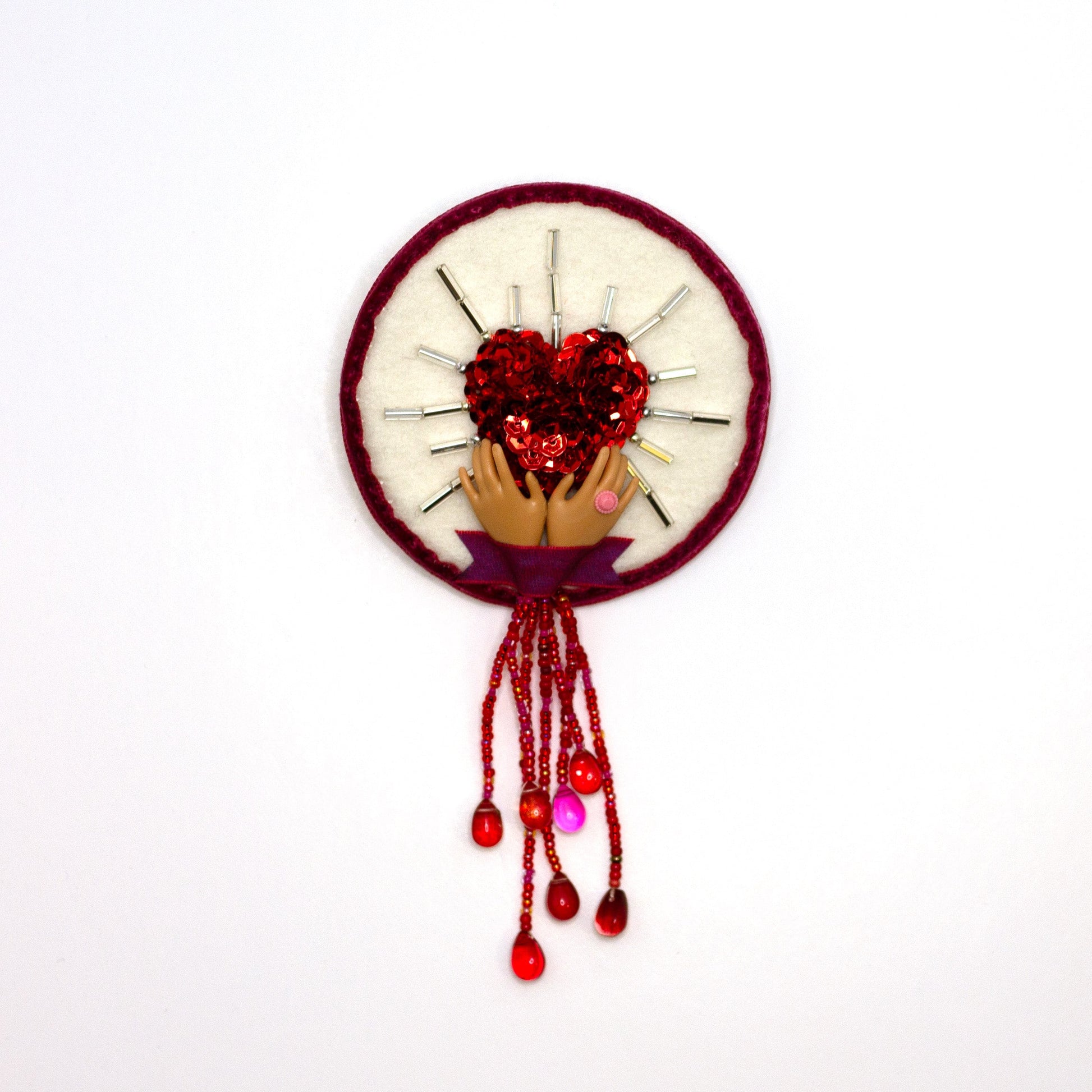 Handmade brooch featuring a sequined heart cradled by doll hands shown on a white background.