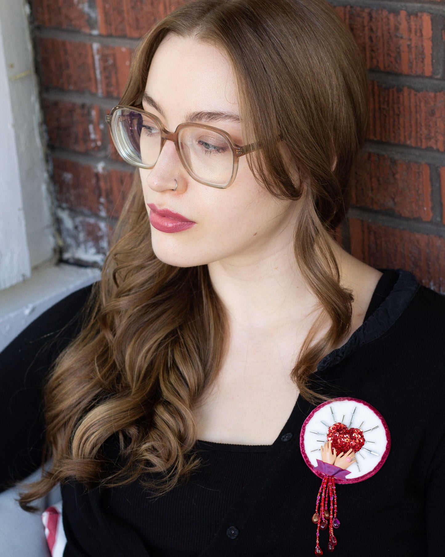 Handmade brooch featuring a sequined heart cradled by doll hands worn on a black sweater by a young woman. 