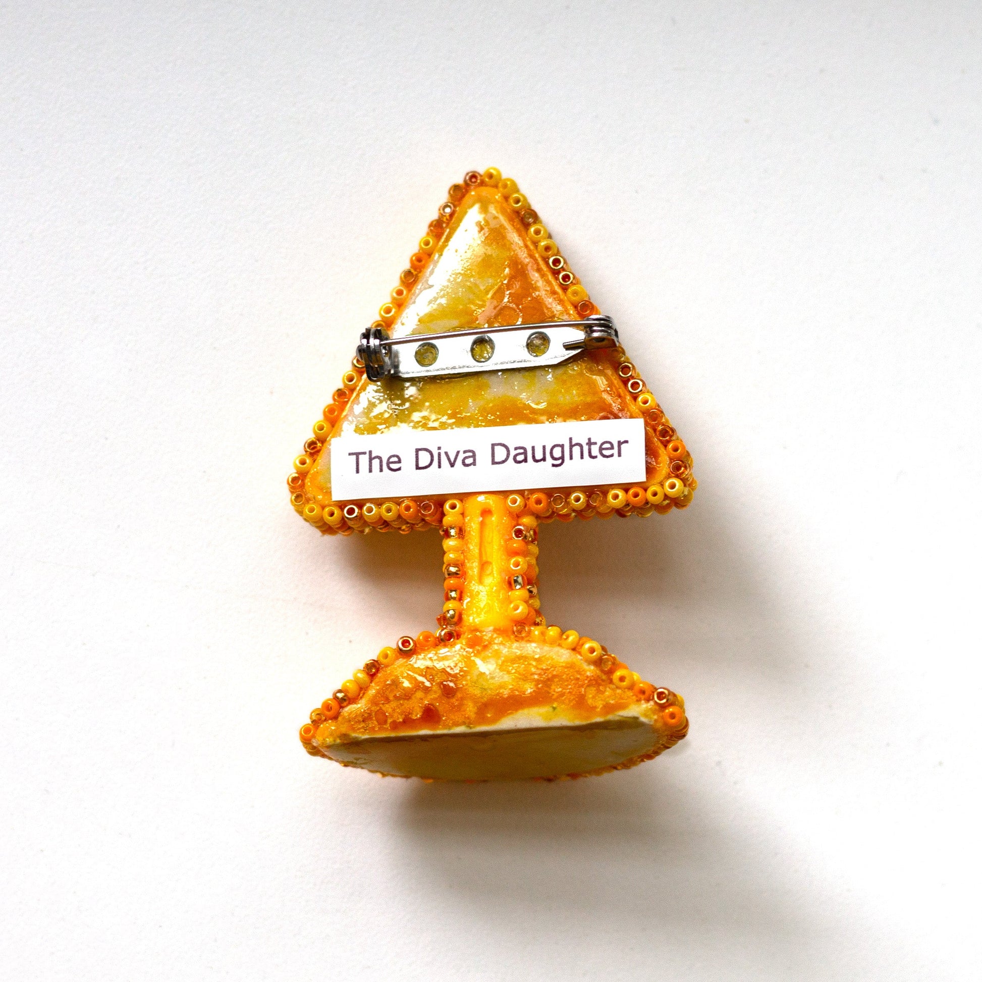 The back of a small triangular gold and orange warning sign pin with a white label reading 'The Diva Daughter' on a plain white background.