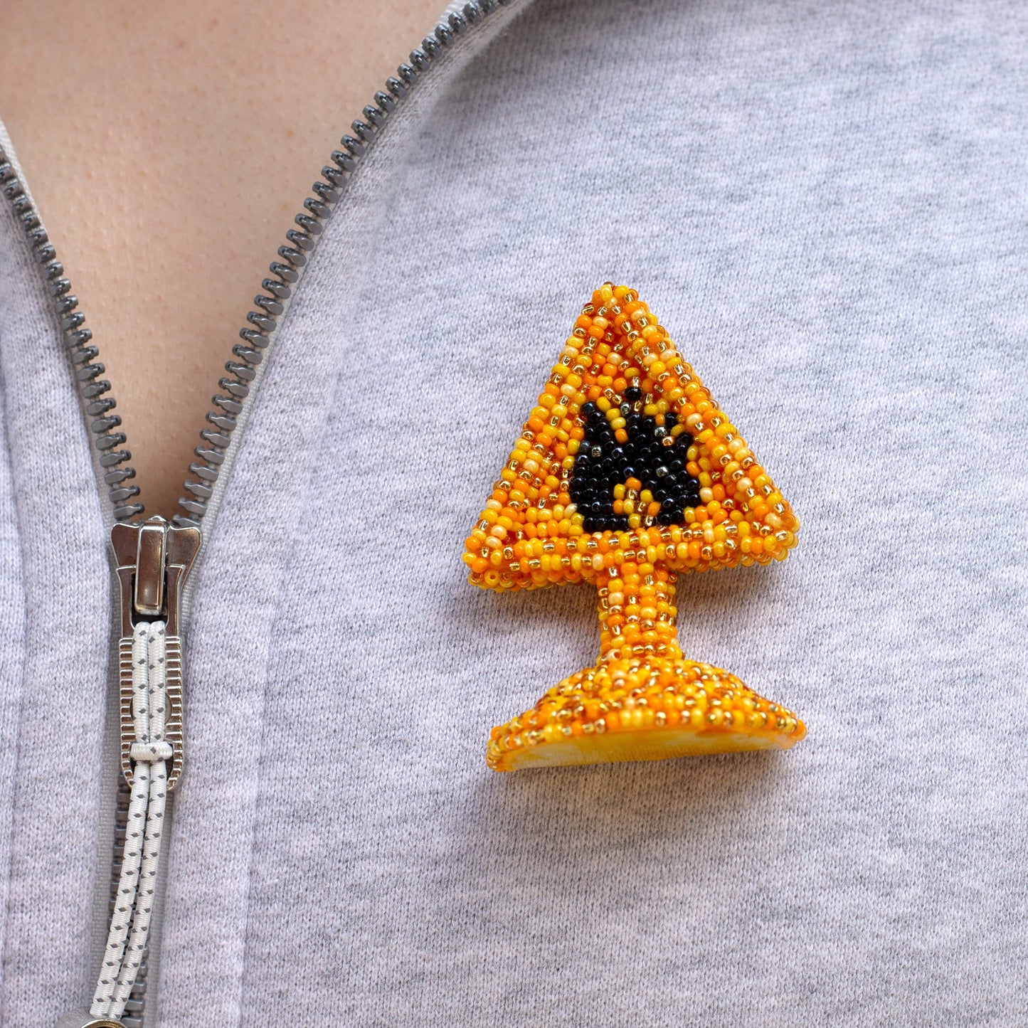 A close-up of a person wearing a gray zip-up hoodie, featuring a small beaded highly flammable pin on the chest. The pin is shaped like a yellow warning sign with a black flame symbol, made from orange and yellow seed beads.