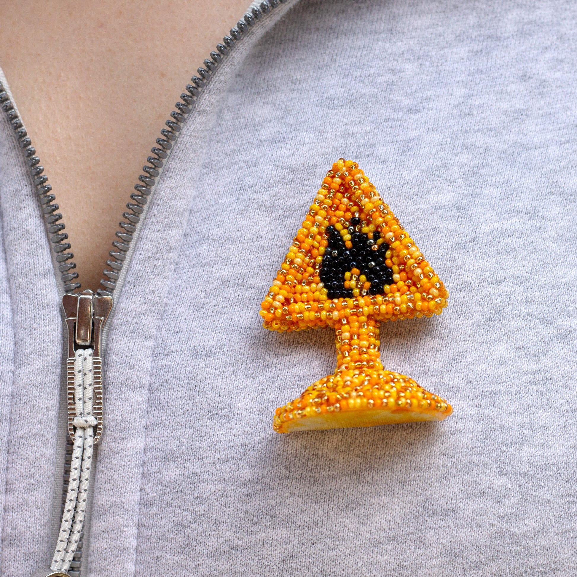 A close-up of a person wearing a gray zip-up hoodie, featuring a small beaded highly flammable pin on the chest. The pin is shaped like a yellow warning sign with a black flame symbol, made from orange and yellow seed beads.