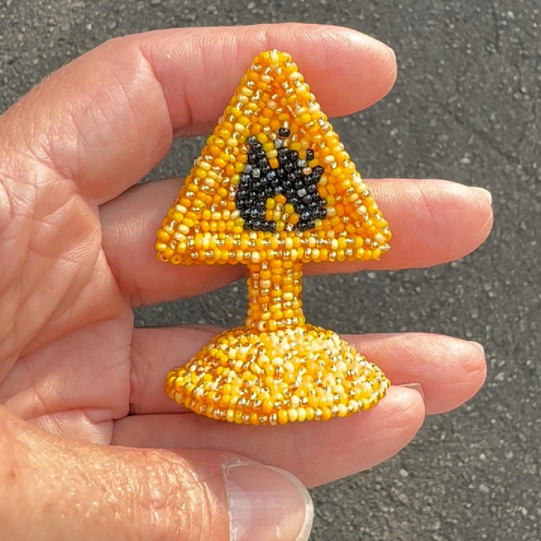 A small beaded highly flammable warning sign brooch in yellow and black, held in a person's hand against an outdoor asphalt background.
