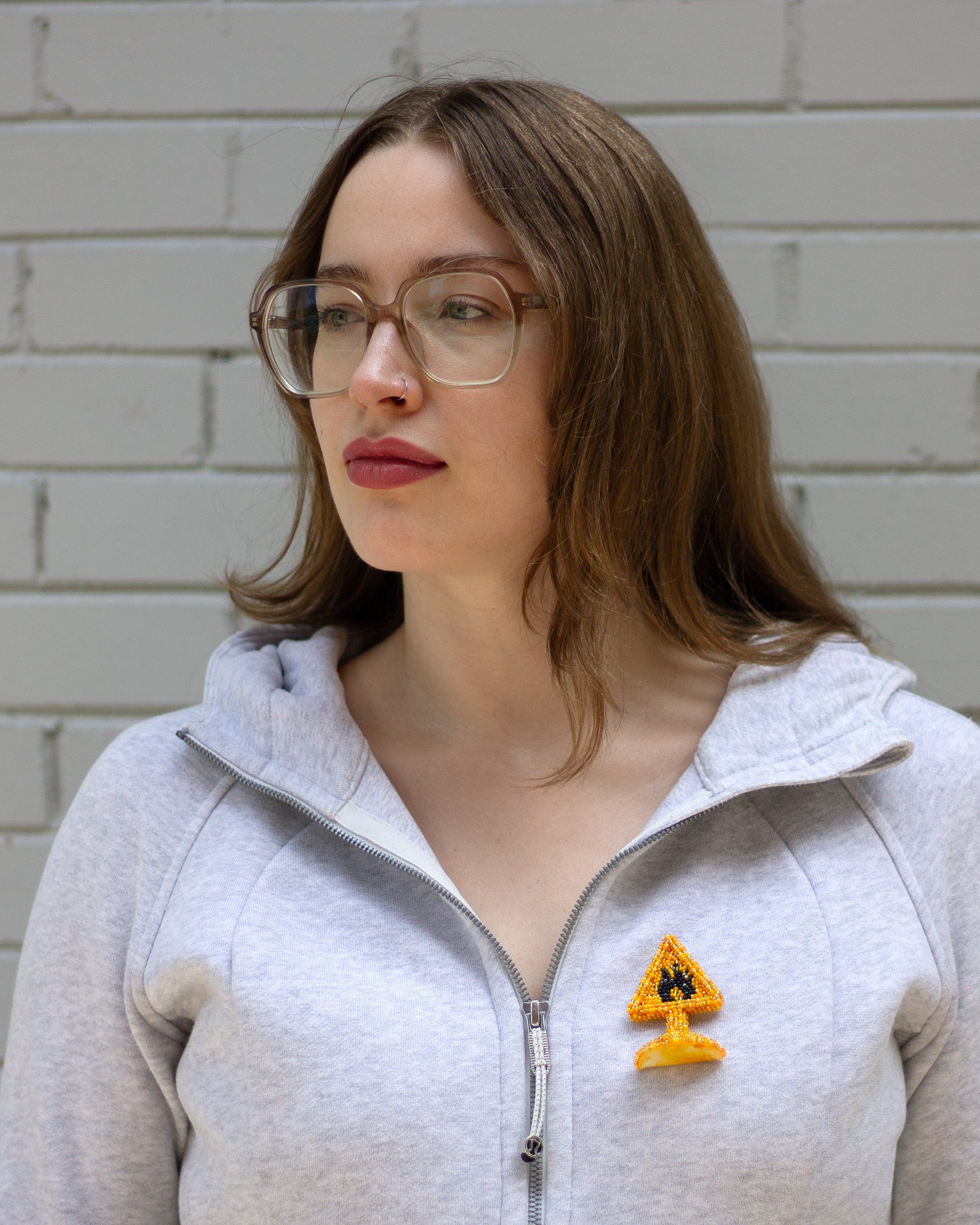 A woman with glasses and brown hair wearing a light gray zip-up hoodie with a small yellow warning sign brooch on the chest, standing against a white brick wall.