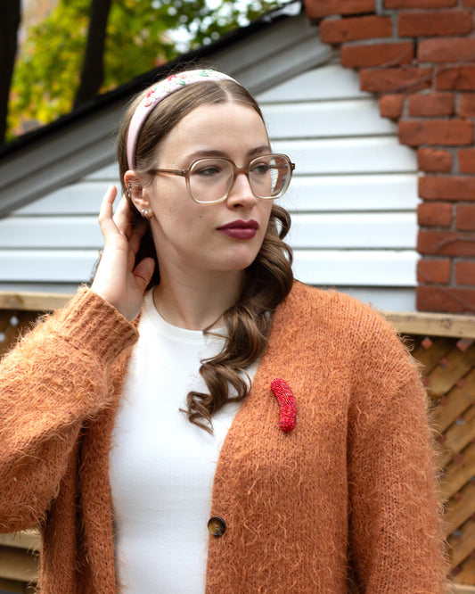 Handmade beaded hot dog brooch worn on an orange cardigan by a young woman.