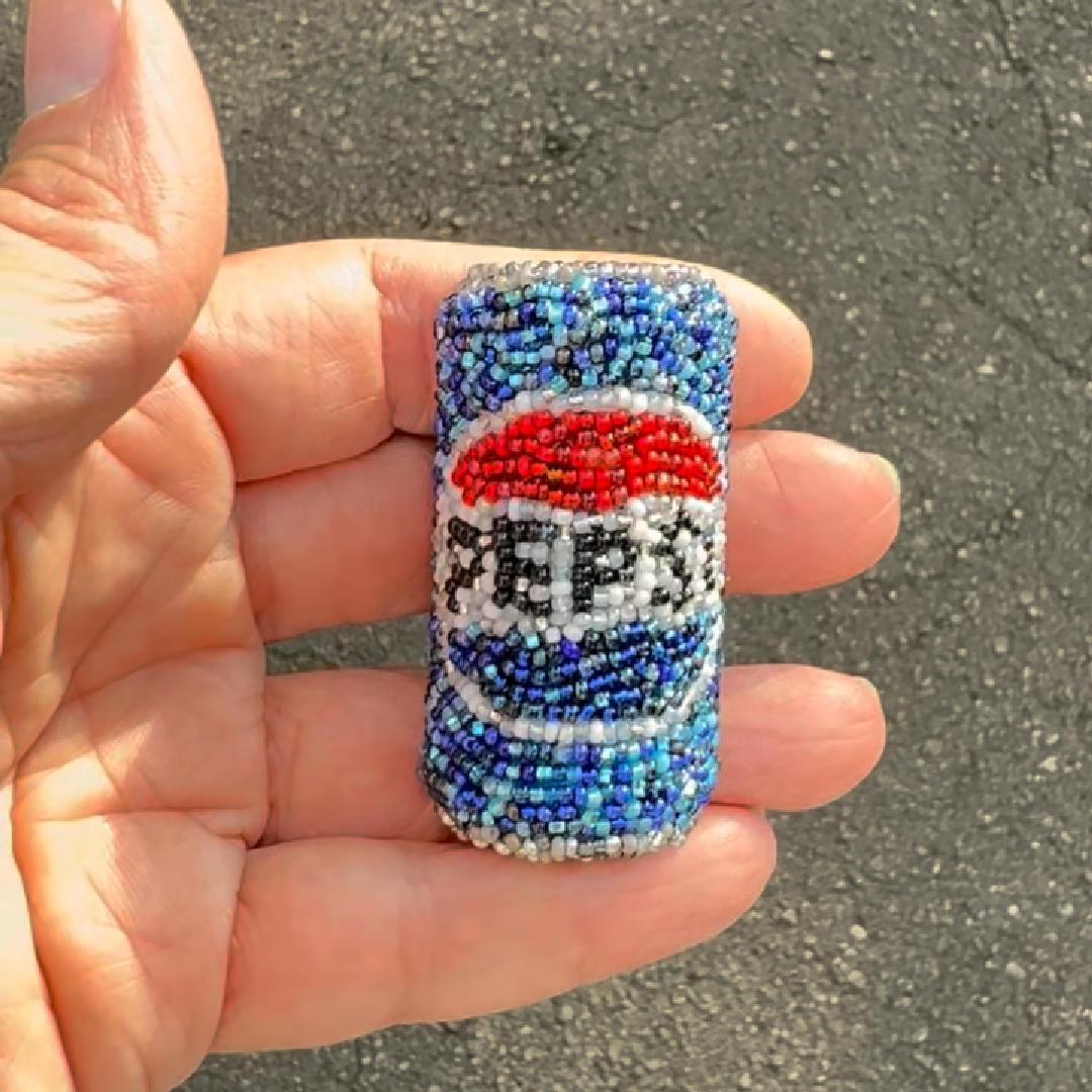 A hand holding a small beaded Pepsi can brooch, featuring blue, white, and red beads with the Pepsi logo.