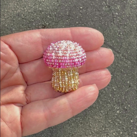 A small pink and beige handmade beaded mushroom brooch held in a hand, taken outside to show off the sparkle.