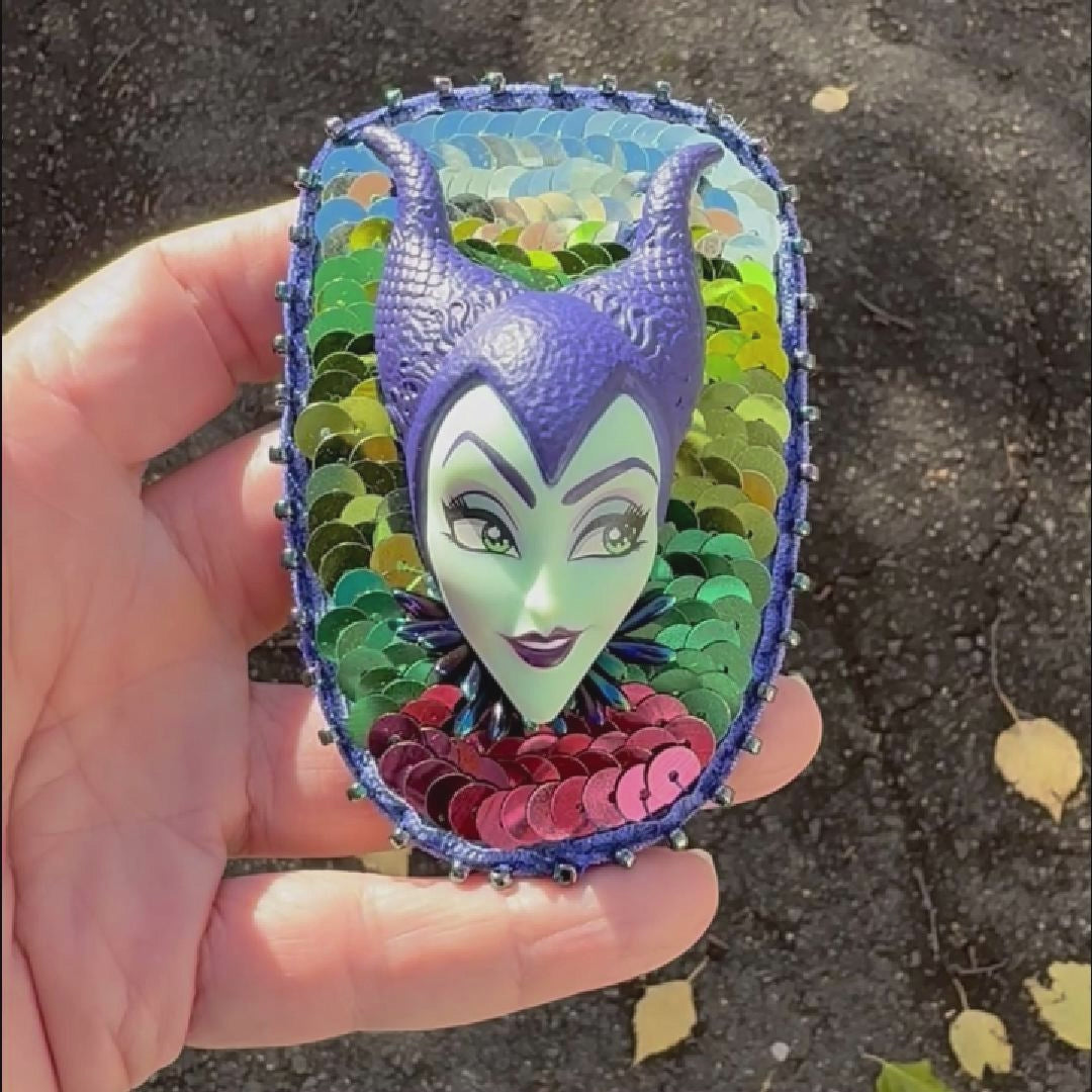 Video of a hand holding a purple and green sequined brooch featuring a Maleficent doll taken outside to show off the spakle.