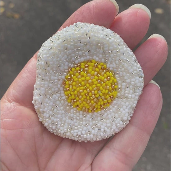 Video showing a hand holding a beaded fried egg brooch taken outside to show off the sparkle.