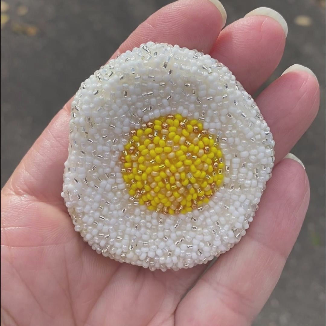 Video showing a hand holding a beaded fried egg brooch taken outside to show off the sparkle.
