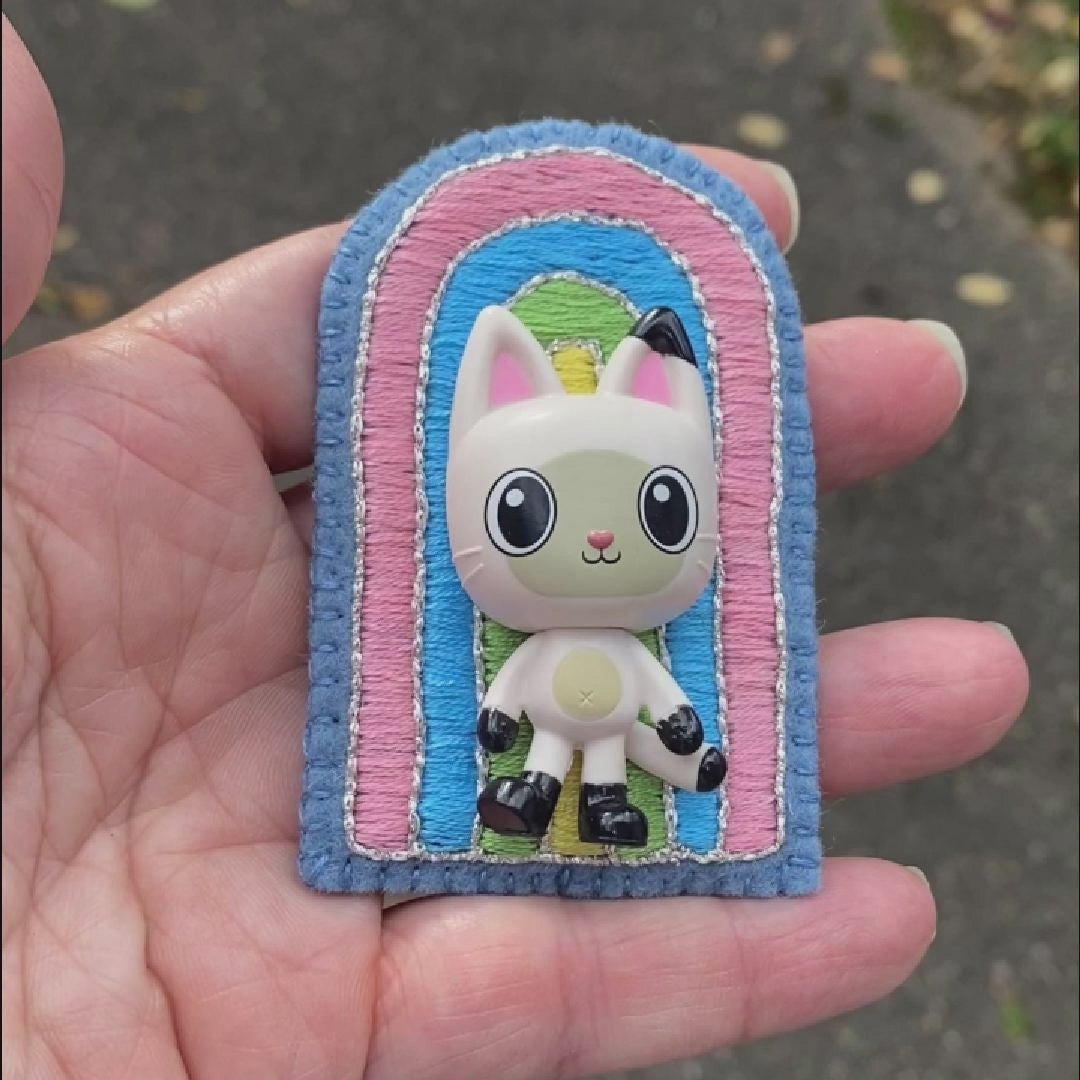 Video of a hand holding an embroidered rainbow brooch featuring Pandy Paws taken outside to show off the sparkle.