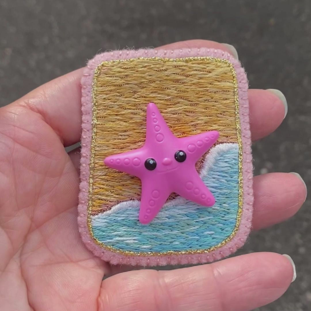 Video of a hand holding an embroidered beach scene brooch featuring a starfish taken outside to show off the sparkle.