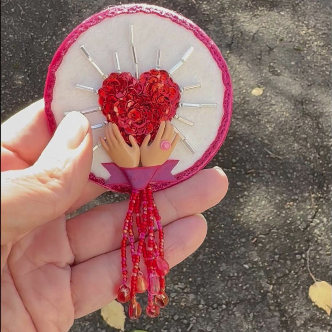 Video of a hand holding a brooch featuring a sequined heart cradled by doll hands taken outside to show off the sparkle.