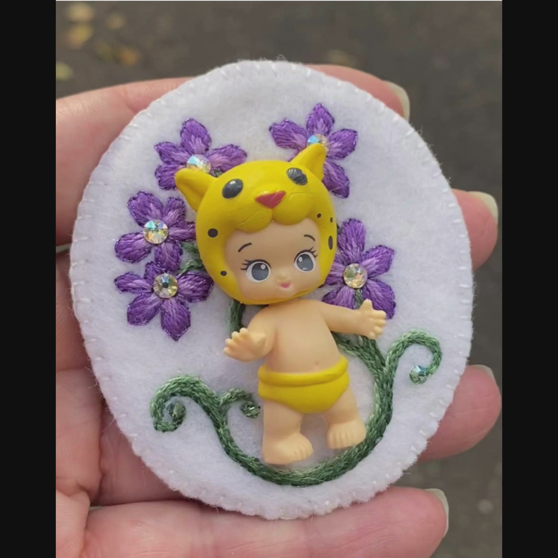 Video of a hand holding an embroidered floral brooch featuring Lippy Twozie taken outside to show off the sparkle.