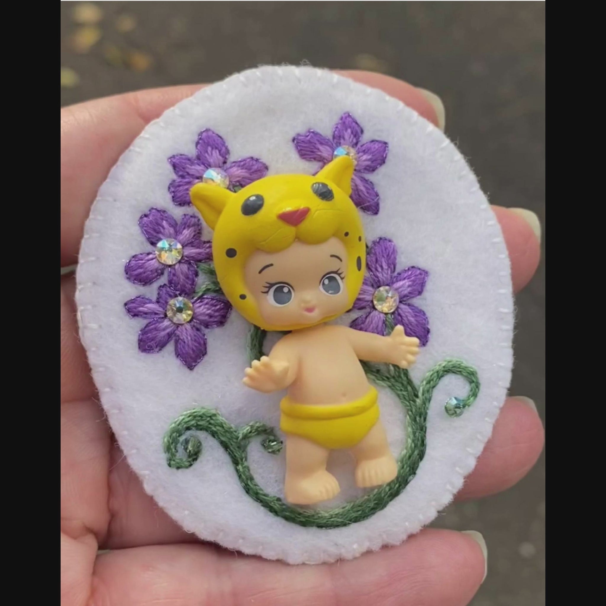 Video of a hand holding an embroidered floral brooch featuring Lippy Twozie taken outside to show off the sparkle.