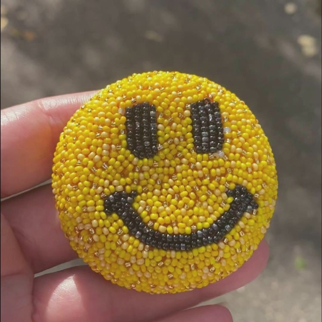 Video of a hand holding a beaded yellow smiley face brooch taken outside to show off the sparkle.