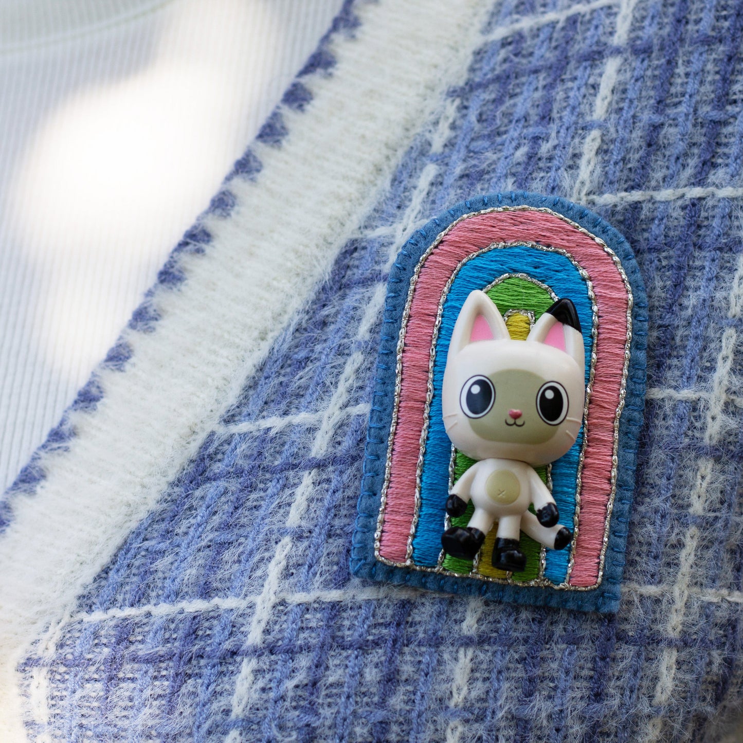 Closeup of a handmade embroidered rainbow brooch featuring Pandy Paws worn on a blue plaid cardigan.