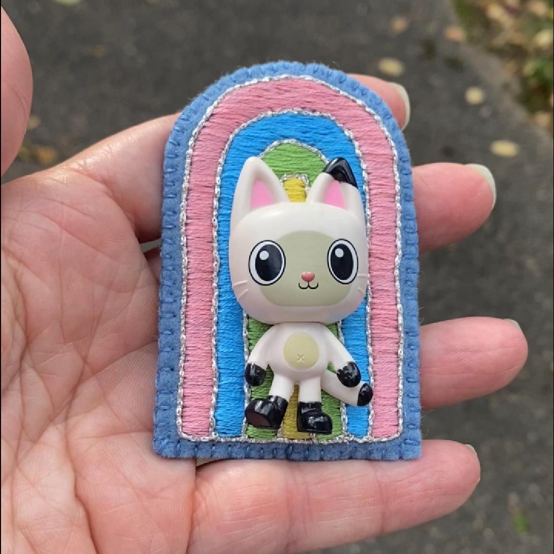 Hand holding an embroidered rainbow brooch featuring Pandy Paws shown in the sunlight.
