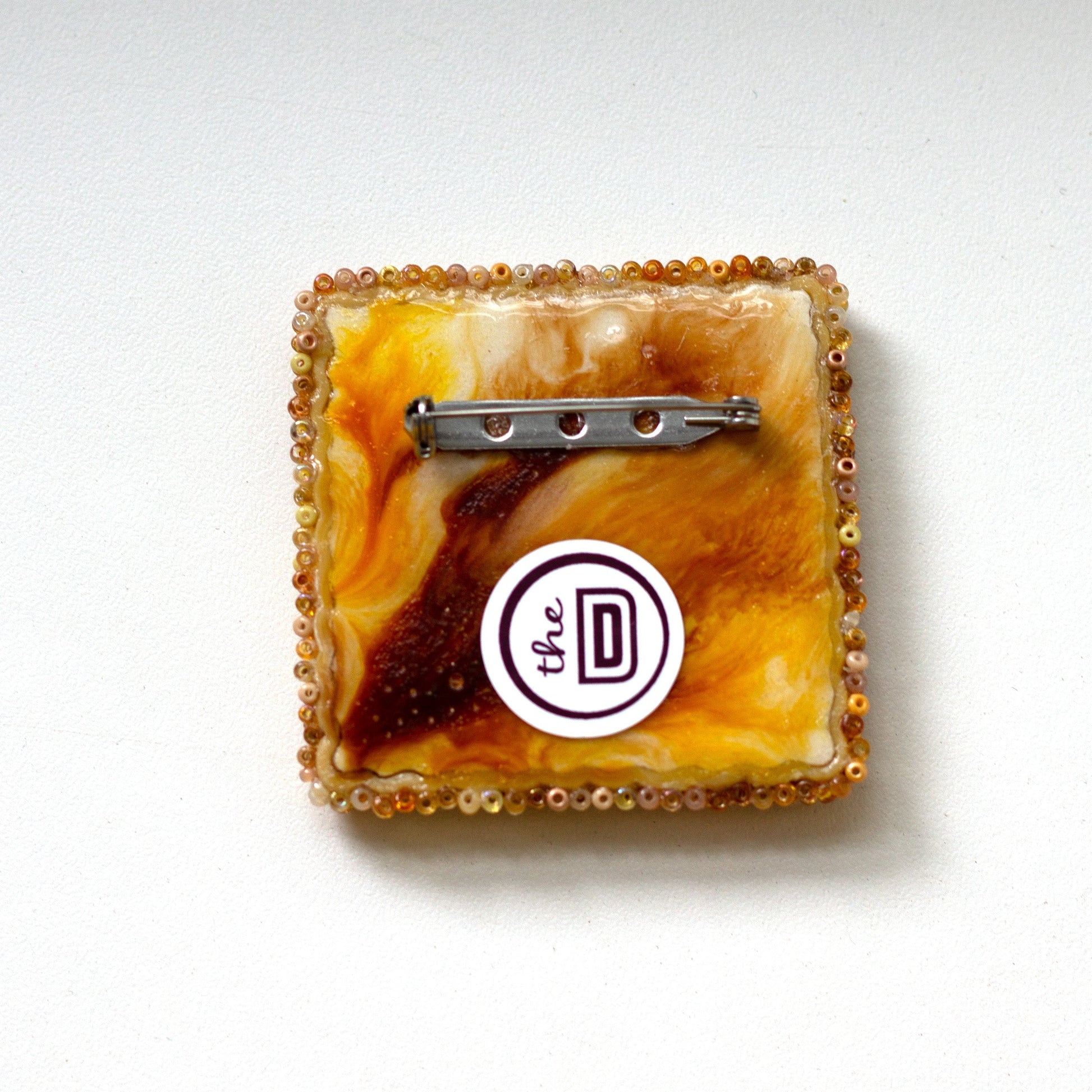 The back of a square beaded cracker brooch with a marbled amber, orange, and brown pattern, featuring a silver-tone metal pin back and a white circular sticker with a black logo.