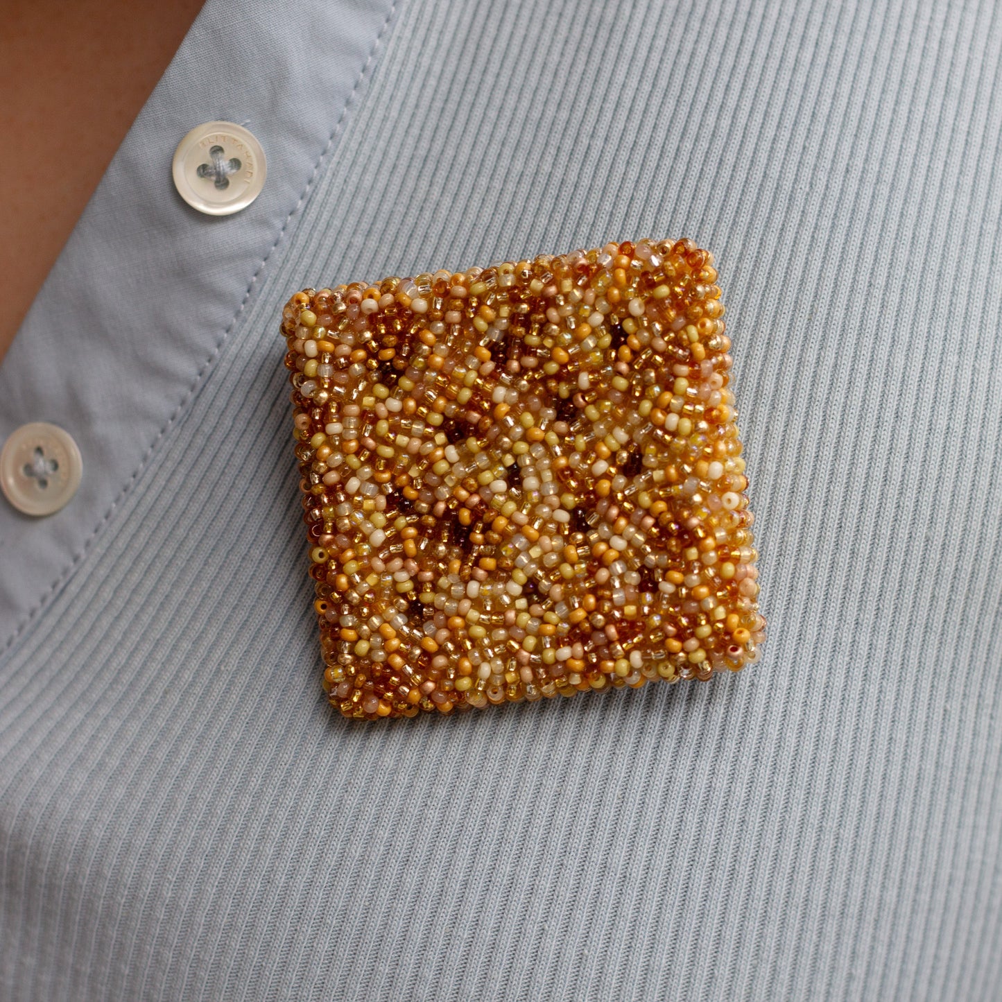A square cracker brooch made of tightly packed amber and cream seed beads, pinned to the chest of a light blue shirt.