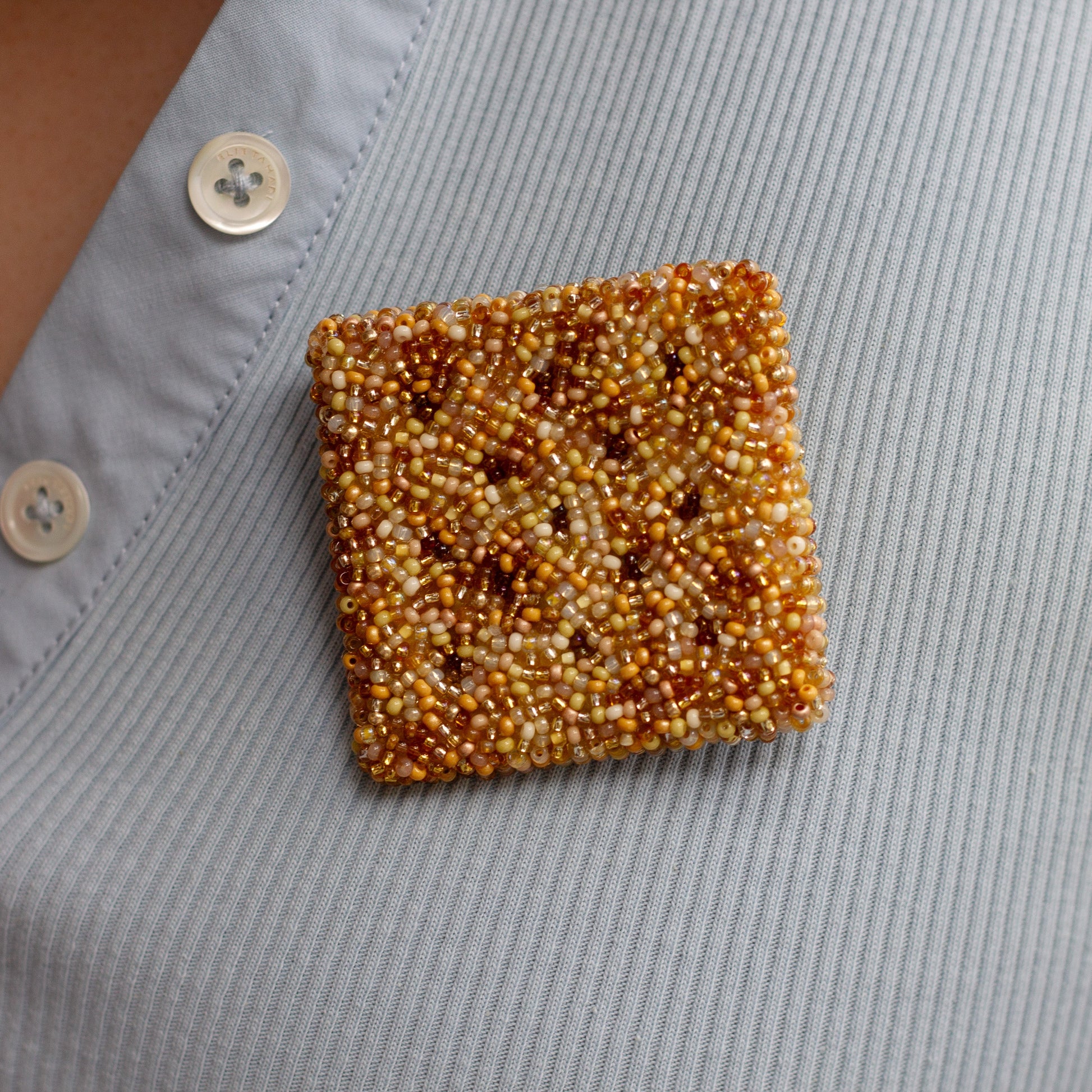 A square cracker brooch made of tightly packed amber and cream seed beads, pinned to the chest of a light blue shirt.